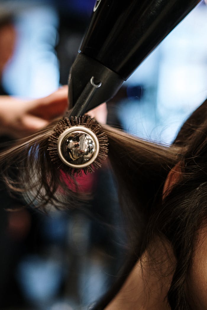 services-01 Close-up of a hairstylist styling hair with a brush and dryer in a salon setting.